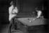 #30201 Historical Black and White Stock Photo of a Woman Leaning On A Cue Stick And Sitting On The Edge Of A Pool Table While Watching Her Friend Play Billiards In 1903 by JVPD